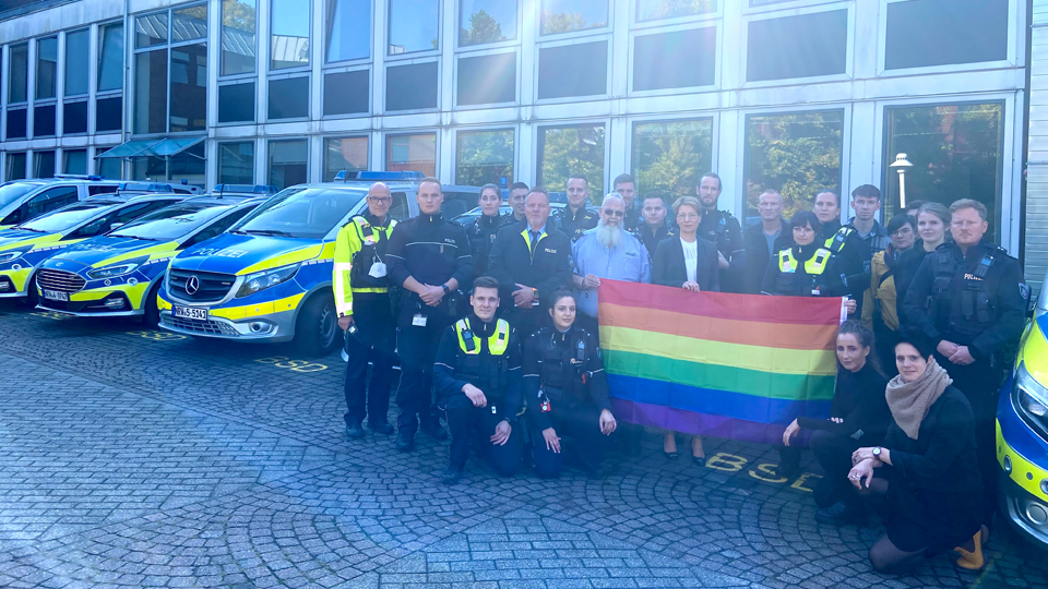 Eine Gruppe von Polizistinnen und Polizisten steht mit Regenbogenflagge vor einer Reihe von Streifenwagen