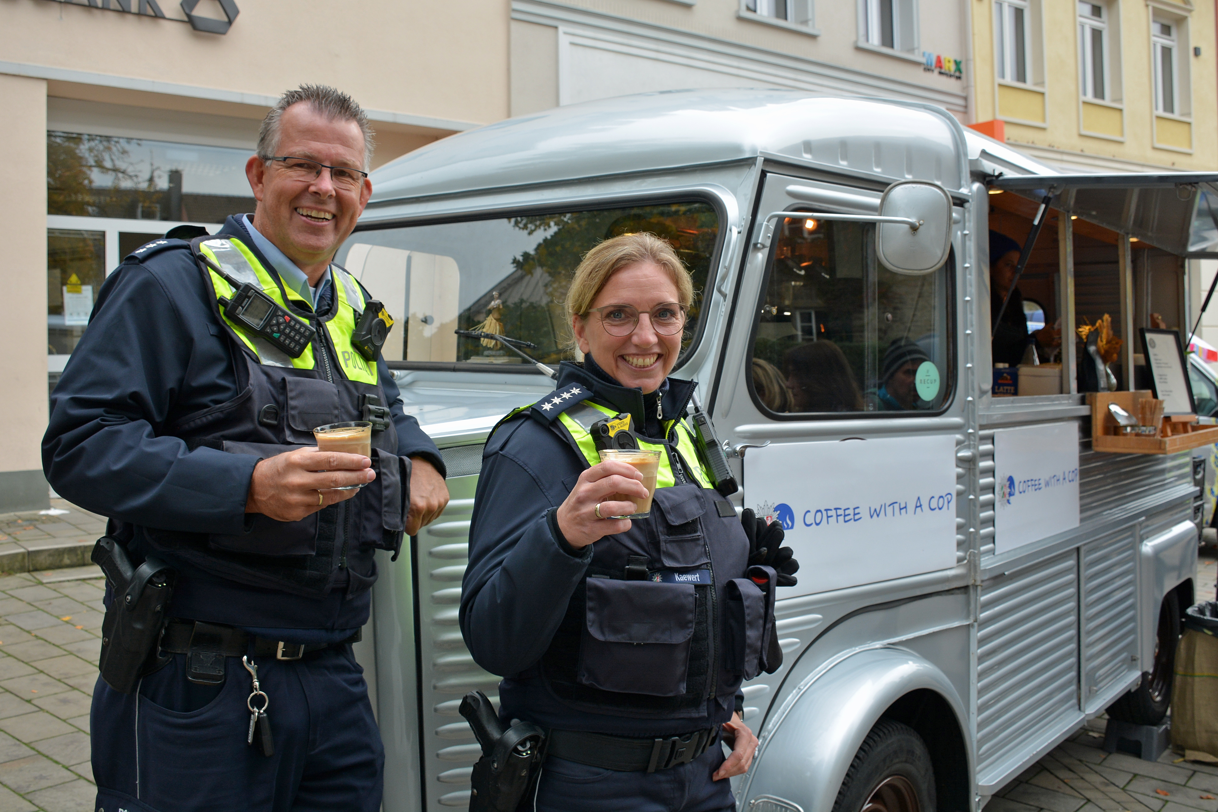 Ein Polizist und eine Polizistin trinken vor einem historischen Kaffee-Mobil einen Kaffee.