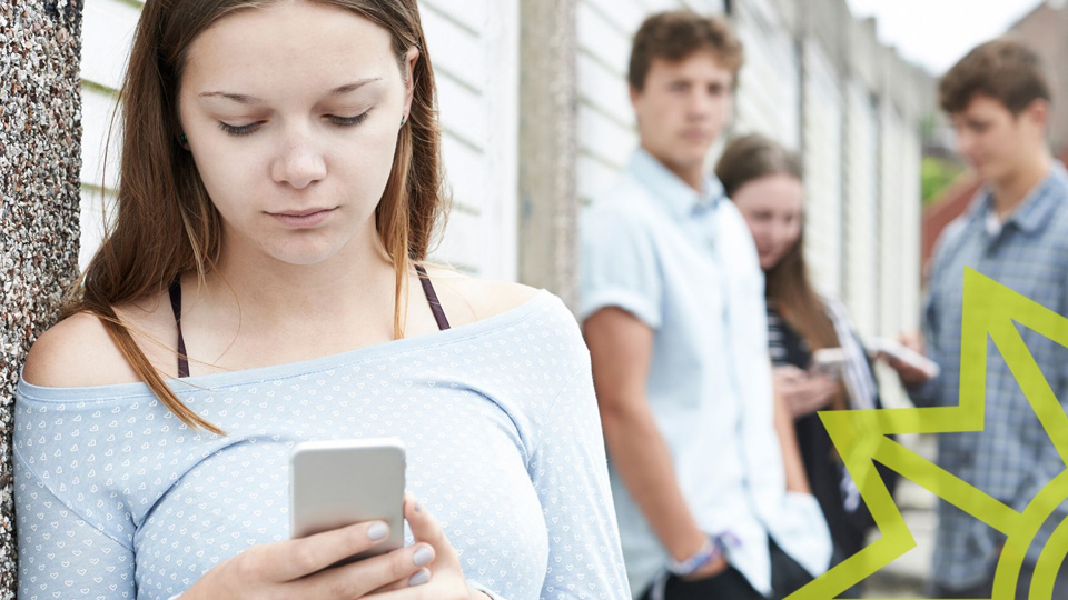 Weiblicher Teenager blickt aufs Handy, im Hintergrund steht eine Gruppe Jugendlicher