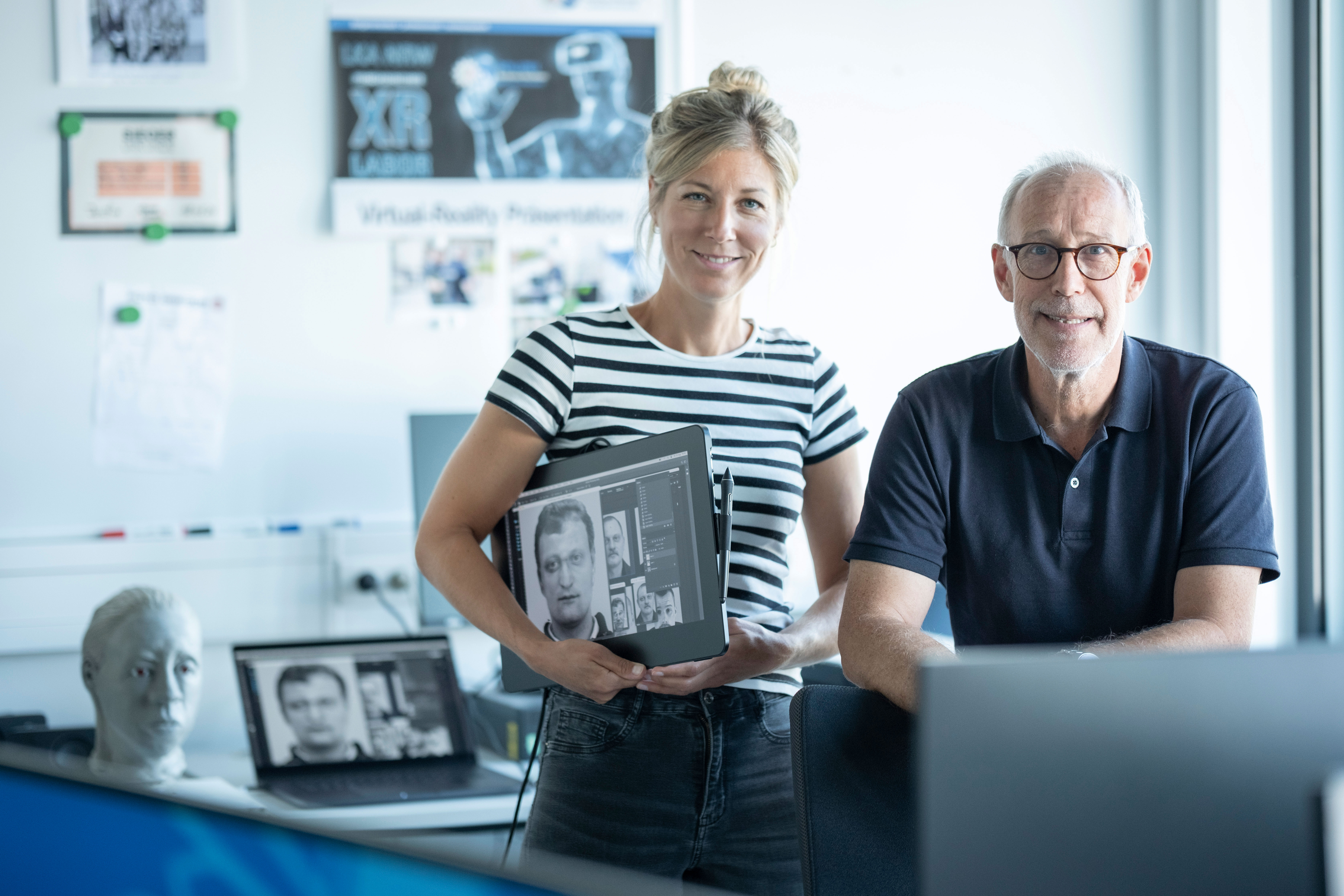 Phantombildzeichner Hanna Mecke und Ingo von Westphal vom LKA NRW befinden sich in einem Büro. Hanna Mecke hält ein Tablet zur Phantombildzeichnung unterm Arm. Beide schauen in die Kamera. Im Hintergrund sind Poster auf einer weißen Wand zu sehen.