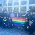 Eine Gruppe von Polizistinnen und Polizisten steht mit Regenbogenflagge vor einer Reihe von Streifenwagen