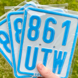 Eine Hand hält drei quadratische blaue Kennzeichen. Eines zeigt "861 UTW".
