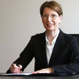 Polizeipräsidentin Alexandra Dorndorf sitzt an ihrem Schreibtisch, lächelt in die Kamera und unterschreibt ein Dokument, links neben ihr steht ein lilafarbener Blumentopf mit einer blühenden Pflanze